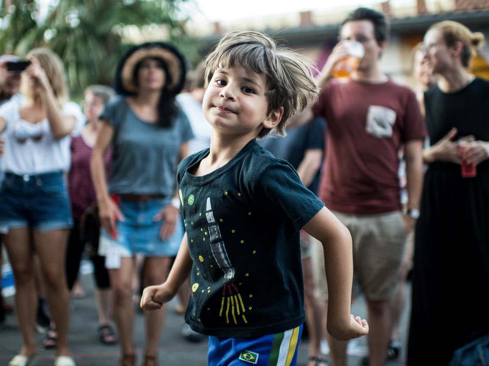 Ein Junge mit einem Raketen-Shirt dreht sich, im Hintergrund verschwommen junge Menschen mit Sommerkleidung. 