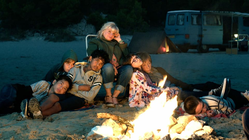 Eine Gruppe von Jugendlichen und eine Frau sitzen am Strand um ein Lagerfeuer herum. 