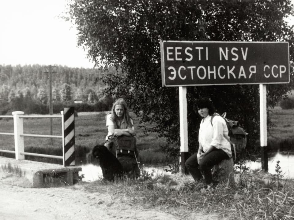 Altes Foto: Zwei Frauen sitzen vor einer Brücke mit Strassenschild.