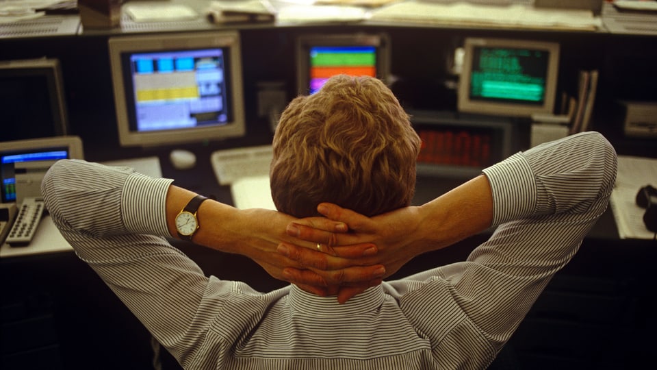 Ein junger Trader sitzt in London vor seinen Bildschirmen mit Börsenkursen, die Hände hinter dem Kopf verschränkt. 