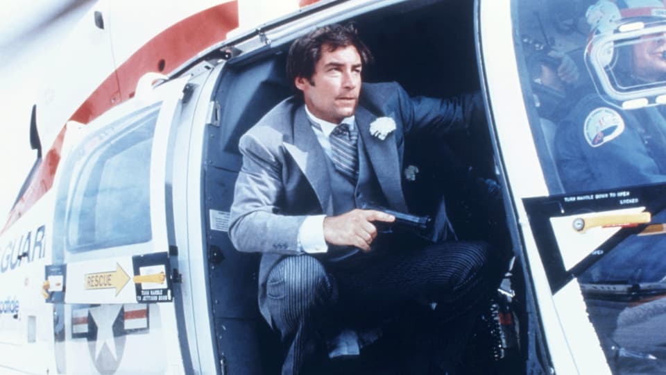 Ein Mann im Anzug und mit einer Pistole in der Hand schaut aus einem Helikopter.