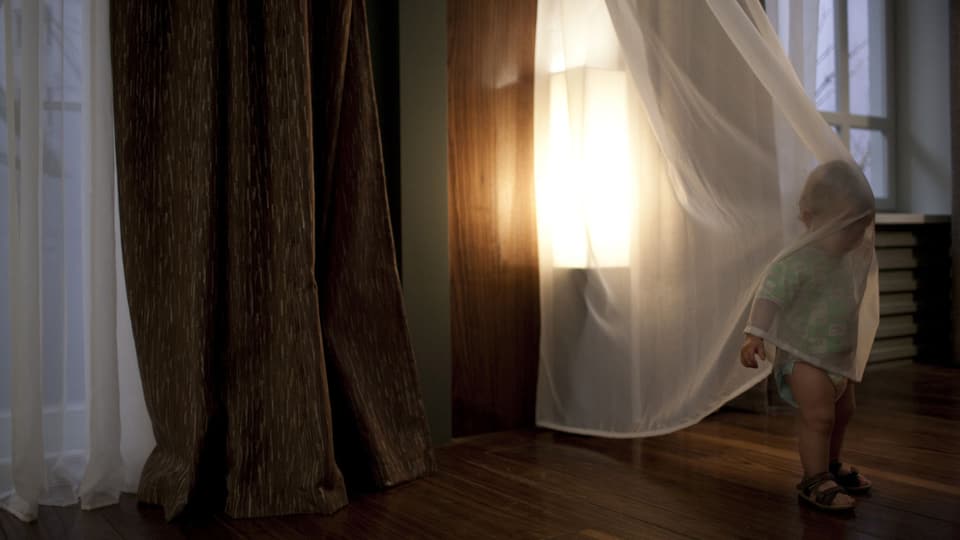 Ein kleines Kind umhüllt sich mit einem durchsichtigen Vorhang, wobei der Kopf und der Oberkörper bedeckt sind. Hinter dem Kind scheint die Sonne durch ein grosses Fenster.