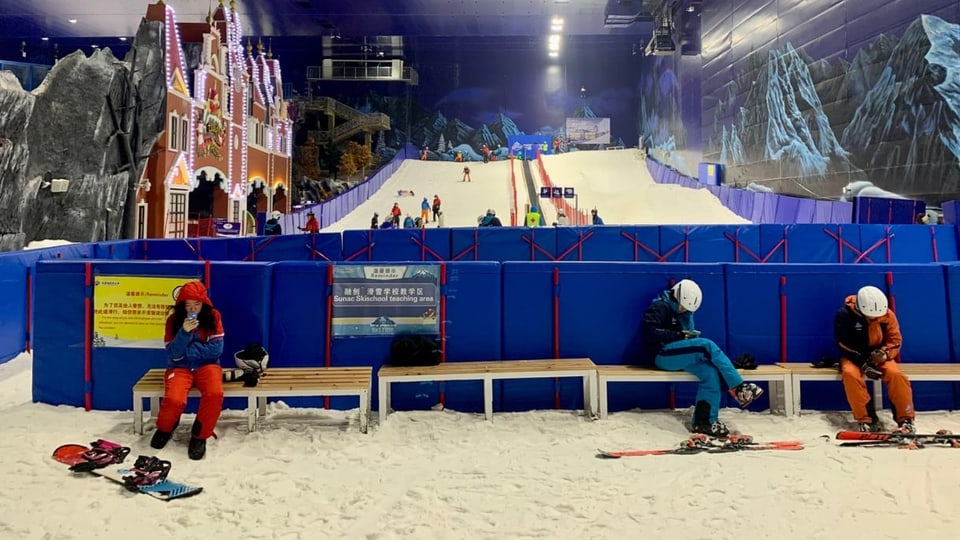 Auf einer Bank sitzen drei Leute in Skikleidung. Sie schauen ins Handy. Hinter ihnen ist die Skipiste.