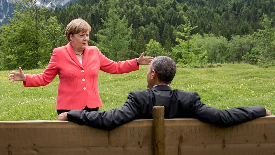 Angela Merkel spricht mit Präsident Barack Obama, der auf einer Bank sitzt.