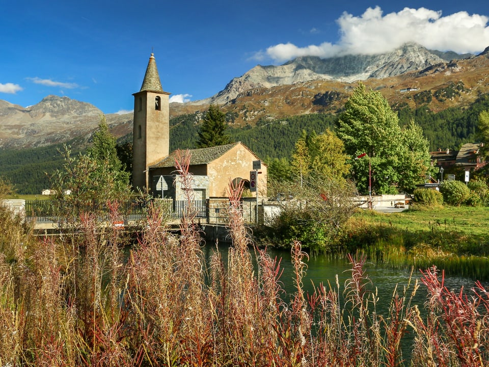 Herbst in einem hohen Alpental mit Kirche. Der Himmel ist blau und die Landschaft grün bis rot. 
