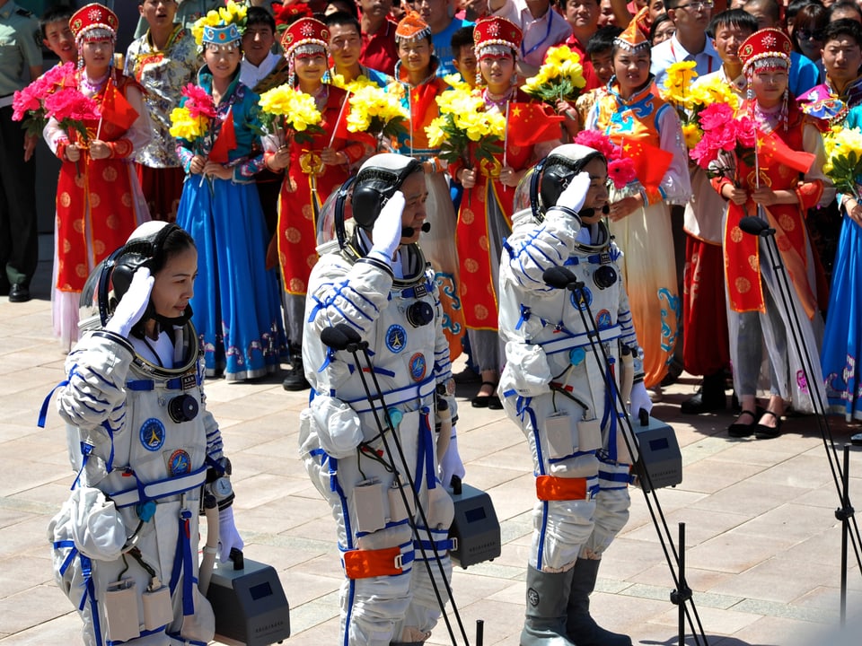Die Astronauten salutieren – dahinter mehrere Frauen in traditionellen Gewändern mit Blumen.