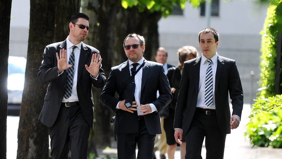 Drei Männer in Anzügen, zwei davon mit Sonnenbrille
