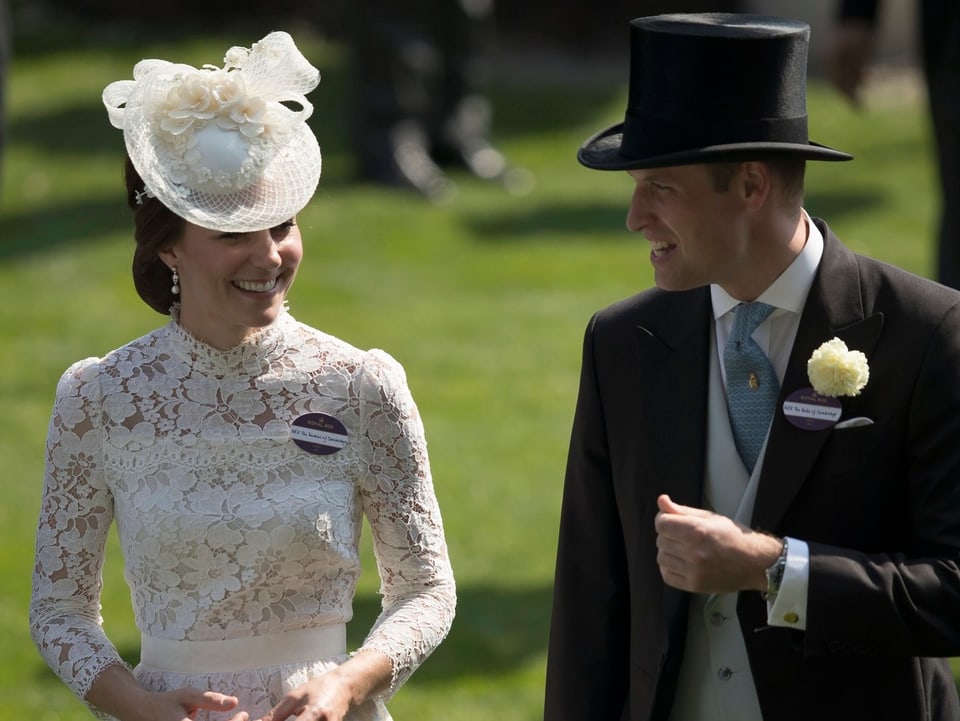 Herzogin Catherine und Prinz William sprechen miteinander. Sie trägt ein weisses Spitzenkleid mit einem Hütchen. Er Zylinder und Frack.