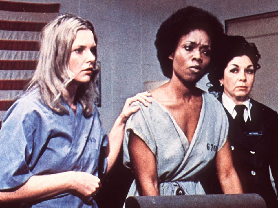 Zwei Frauen im Häftlingsanzug, daneben eine Frau in Uniform. 