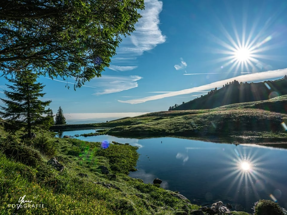 Sonnenaufgang am nur leicht bewölkten blauen Himmel. Die Sonne spiegelt sich in einem Bergesee.