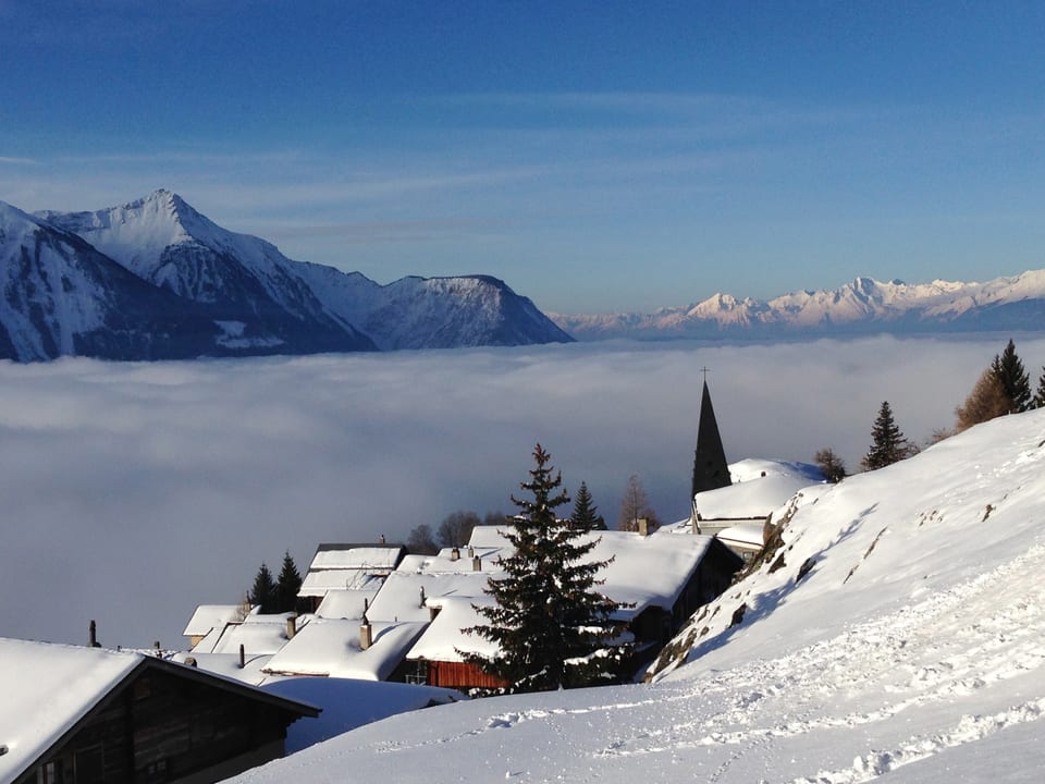 Winterliches Dorf am Berghang an der Sonne. Im Tal liegt Nebel. Darüber scheint die Sonne vom blauen Himmel. 