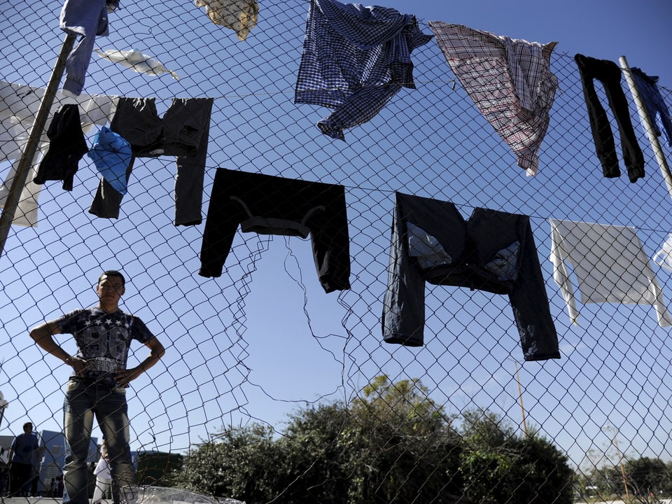 Ein Flüchtling steht vor einem ramponiertem Zaun, auf dem zahlreiche Kleidungsstücke zum Trocknen hängen. 