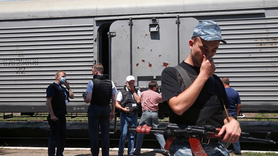 Mann mit Gewehr hält sich die Nase zu. Dahinter ist eine Gruppe Personen vor einem Eisenbahnkühlwagen zu sehen.
