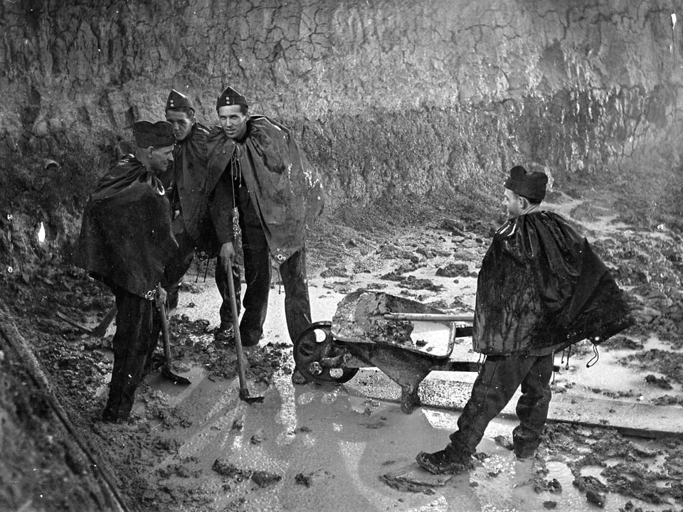 Vier Soldaten mit Regenpellerine stehen in einer Dreckhalde. In der Mitte steht eine Schubkarre. Es regnet.