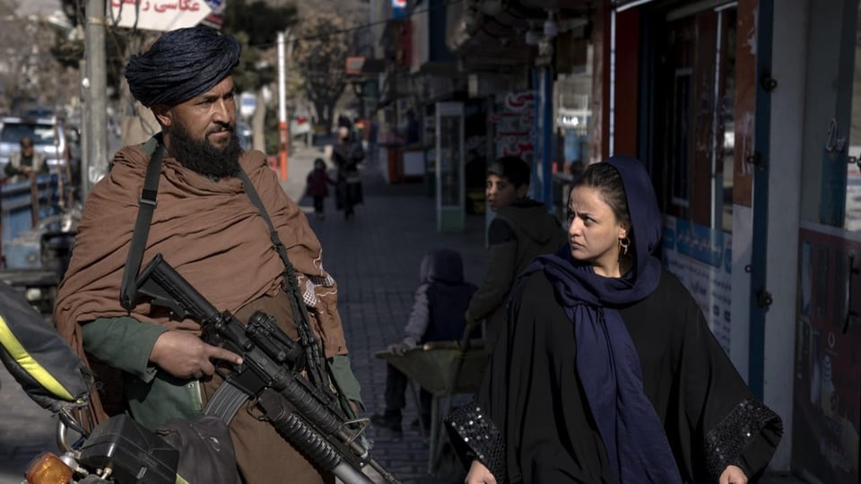 Frau läuft neben Mann mit Maschinengewehr und blick ihn an. Er ist ein Taliban.