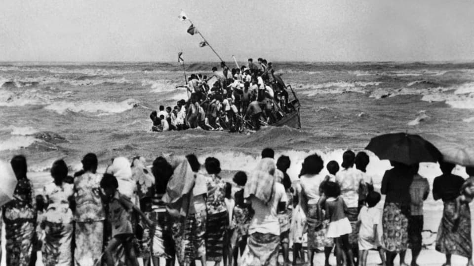 Schwarzweissfoto: Menschen an einem Strand schauen auf ein kleines Boot im Meer mit unzähligen Menschen