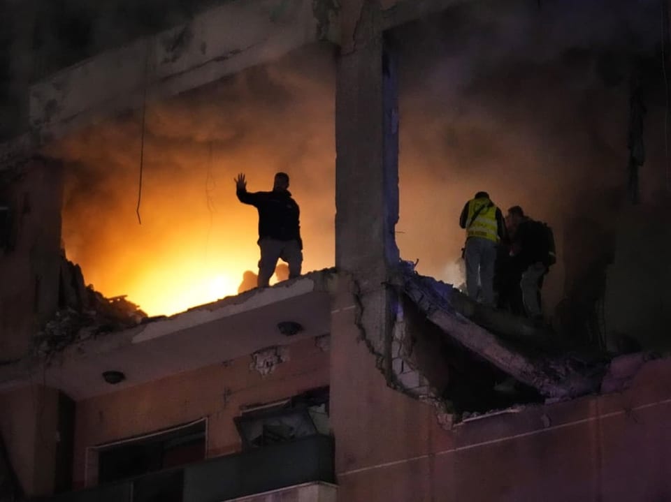 Männer stehen in einem Geschoss eines Gebäudes. Die Fassade fehlt. Im Hintergrund scheint es zu brennen.