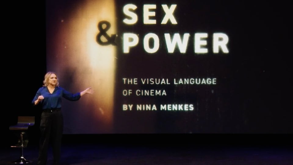 The speaker stands in front of a large screen with the words “Sex and Power” written on it.