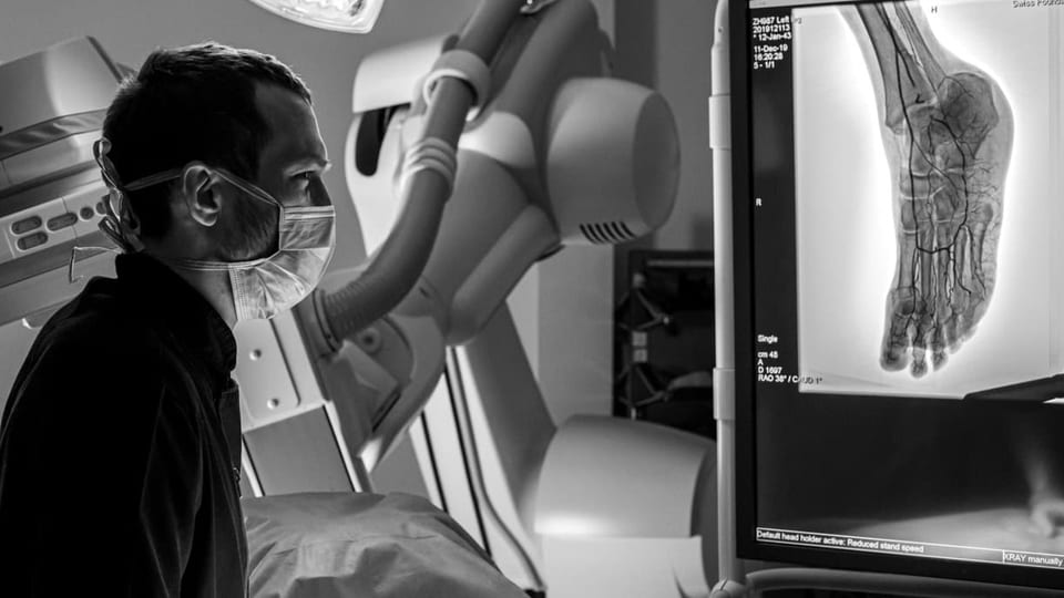 Ein Mann mit Maske steht vor einem Bildschirm an dem man einen Fuss mit Blutgefässen sieht, ähnlich wie ein Röntgenbild