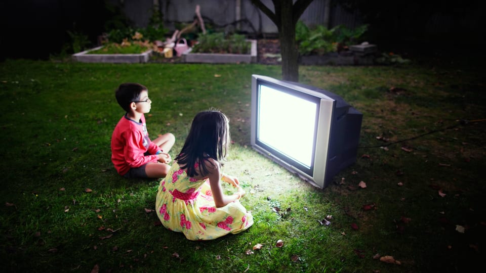 Zwei Kinder sitzen vor einem Fernseher auf einer Wiese
