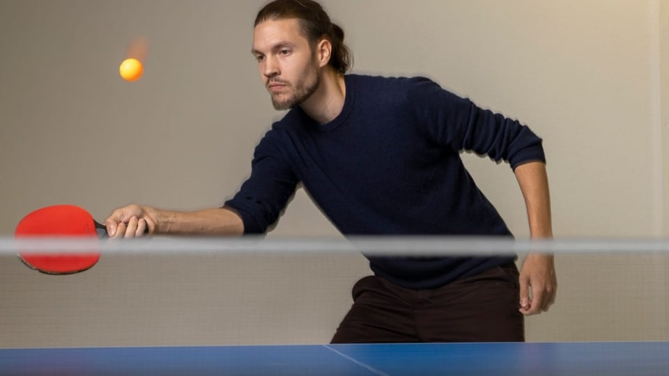 junger Mann mit dunklem Pferdeschwanz hinter Pingpongtisch, hält Schläger, schaut auf Ball in der Luft.