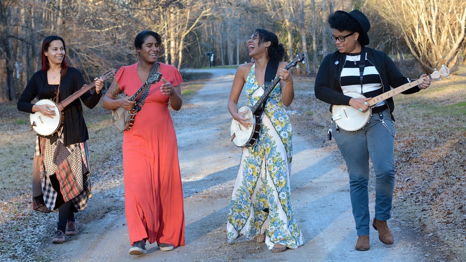 Vier Frauen in bunten Kleidern, jede mit einem Banjo, spielend, auf einem Feldweg