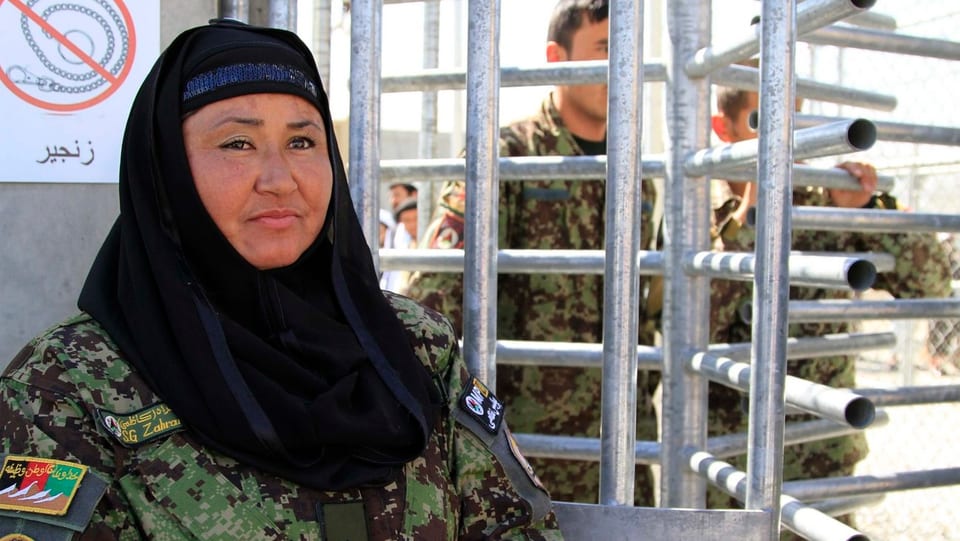 Frau in Militäruniform mit einem schwarzen Kopftuch steht vor einem Gitter-Drehtor.
