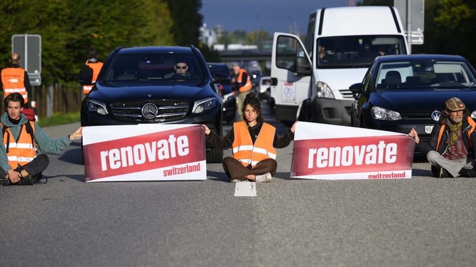 Personan sitzen auf einer Fahrbahn und halten ein Plakat in der Hand.