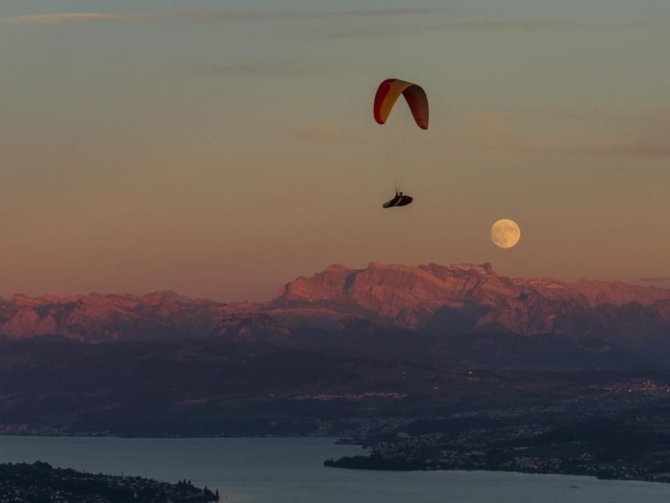 Gleitschirm am Abendhimmel mit Mond am Horizont über den Alpen, unten Zürichsee im Schatten. 