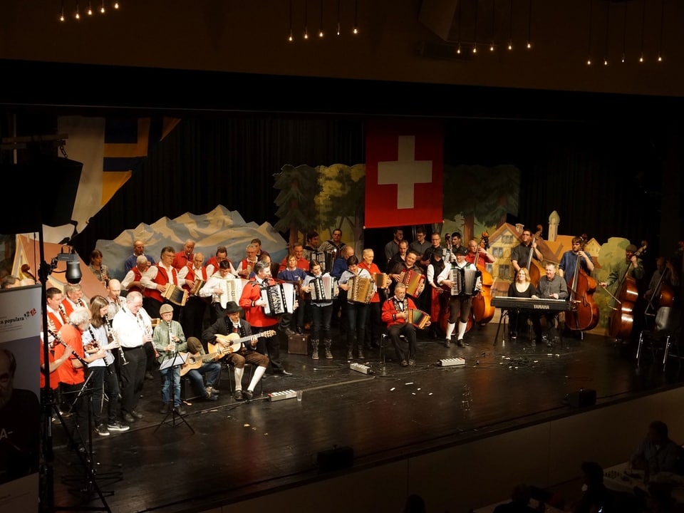 Eine grosse Gruppe von Musikantinnen und Musikanten mit ihren Instrumenten auf einer Bühne.