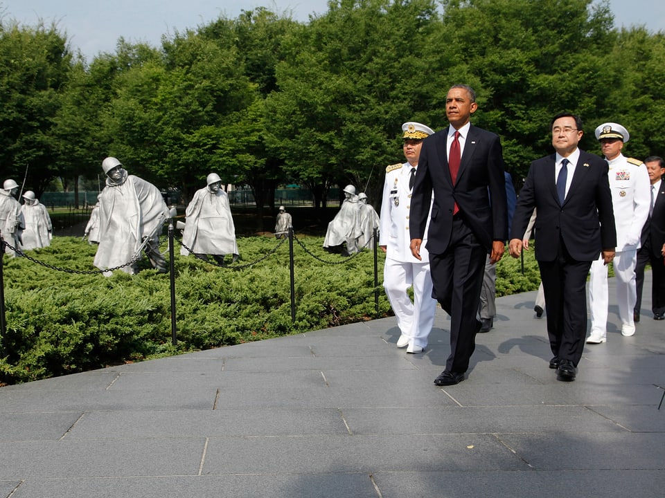 Im Hintergrund: Statuen aus Stahl, die Soldaten repräsentieren. Vorne durch läuft US-Präsident Barack Obama mit einer Delegation.