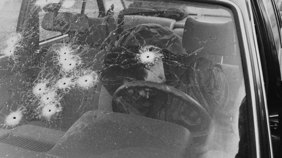Fensterscheibe eines Autos, das von zahlreichen Schüssen durchsiebt ist. Dahinter sitzt der Fahrer des Autos tot.