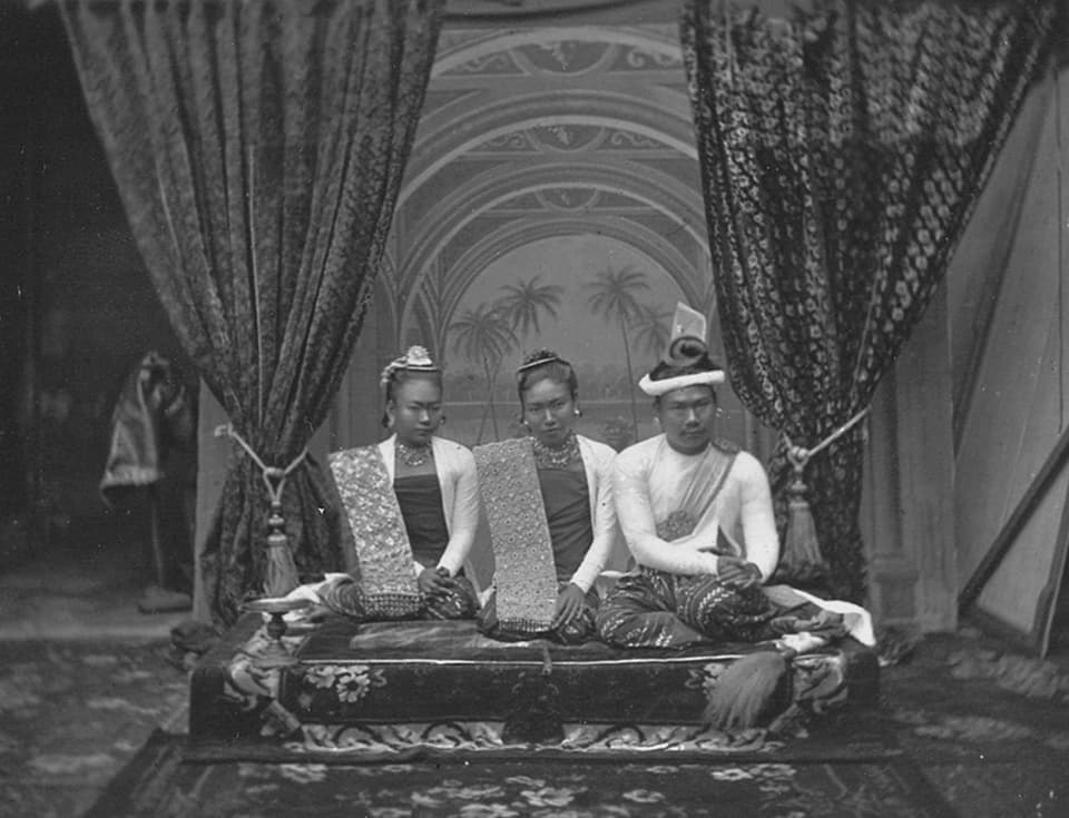 Foto von zwei Frauen und dem burmesischen König auf einer Erhöhung, hinten Vorhänge.