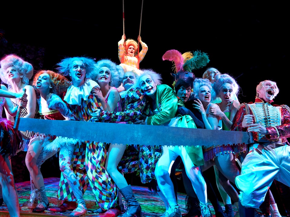 Als Zirkusartisten und Clowns verkleidete Schauspieler ziehen an einer Riesensäge.