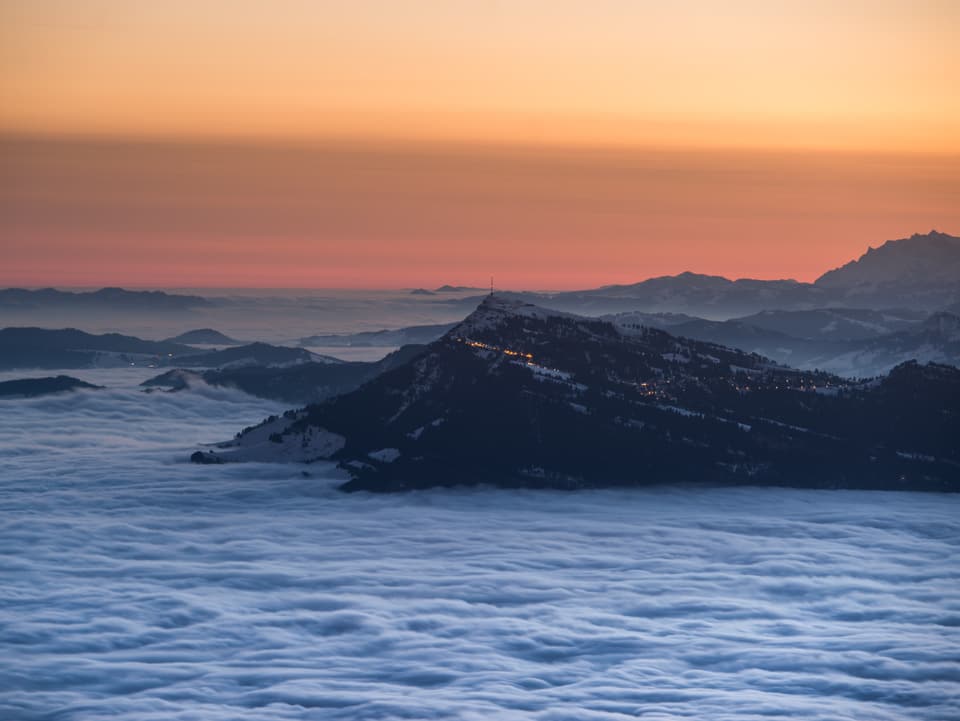 Die Rigi, ein spitzer Berg, ragt aus dem Nebelmeer. Dahinter der baldige Sonnenaufgang.
