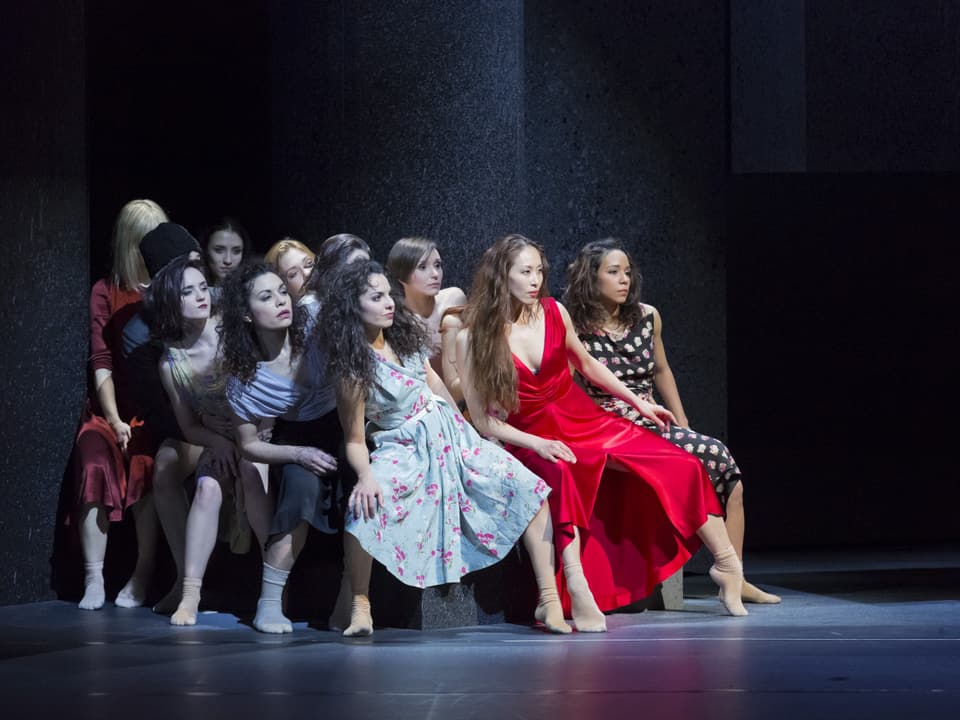 Eine Gruppe von Frauen tanzt auf einer Theaterbühne. Sie formieren sich in einer Gruppe und tragen knielange, unterschiedlich farbene Kleider.