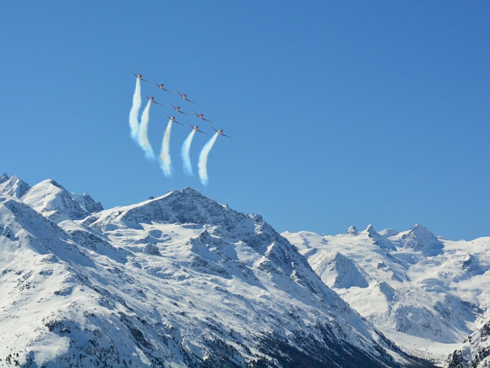 Der Himmel ist wolkenlos und die verschneiten Berge strahlen. Am Himmel ziehen 9 Militärflugzeuge 5 Rauchschwaden hinter sich her.