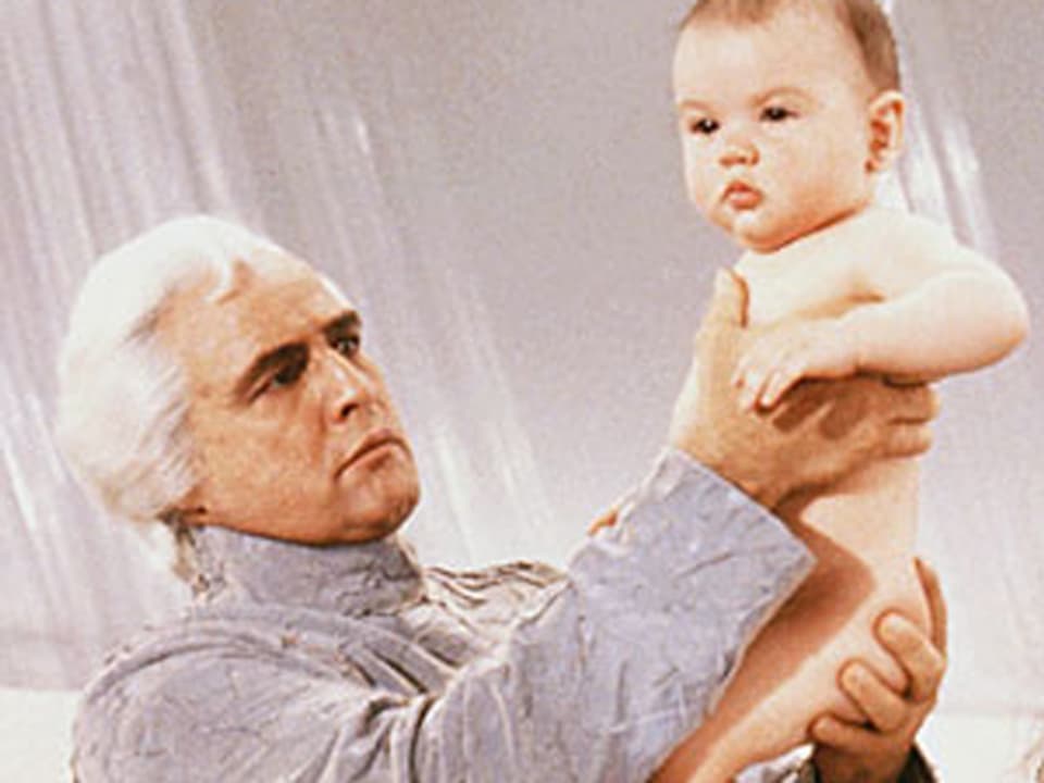 Ein älterer Mann mit weissem Haar hält ein nacktes Baby in seinen Händen.
