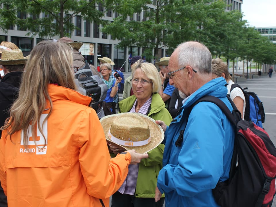 Frau in oranger Jacke übergibt einem Wanderpaar einen Strohhut.