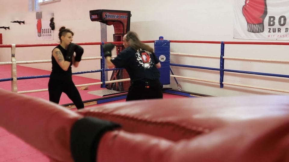 Boxring, links boxt eine junge Frau in schwarz, rechts ein älterer Mann mit langen Haaren, auch schwarz gekleidet.