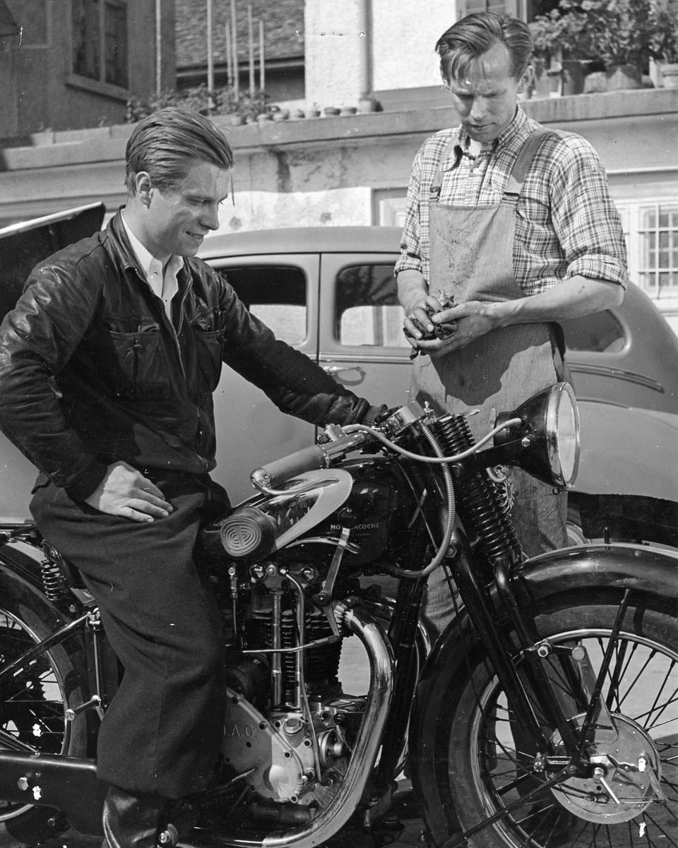 Ein Mann sitzt stolz auf seinem Motorrad. Ein zweiter Mann steht daneben. Beide betrachten das Motorrad.