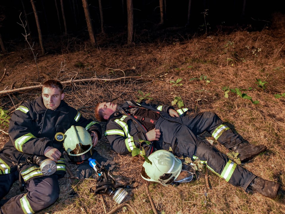 Zwei Feuerwehr-Männer haben sich auf den Waldboden gelegt, um sich auszuruhen. 