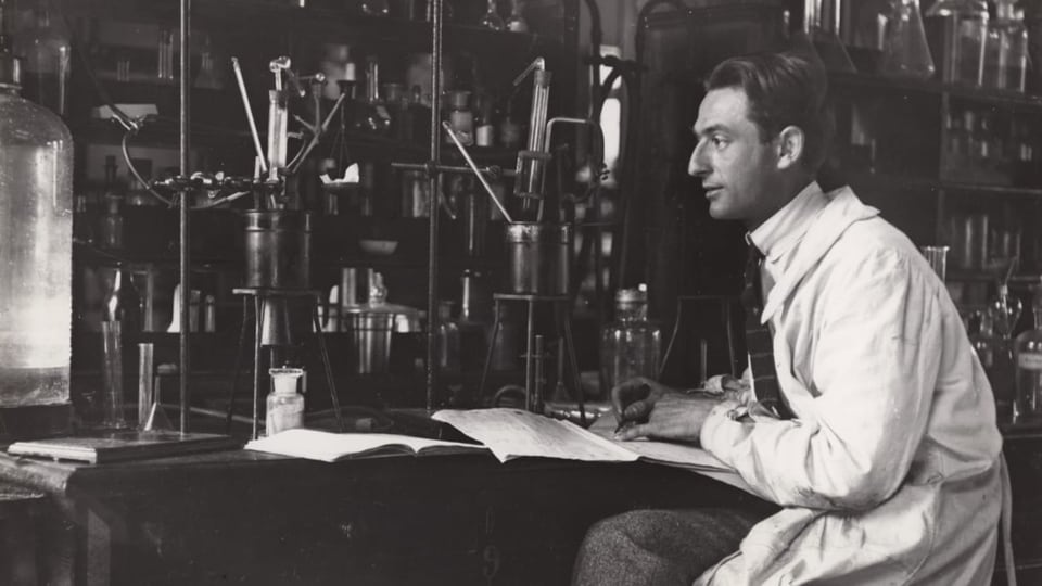 Ein Mann in weissem Labor-Kittel sitzt in einem Labor und hat chemische Geräte vor sich auf dem Tisch stehen.