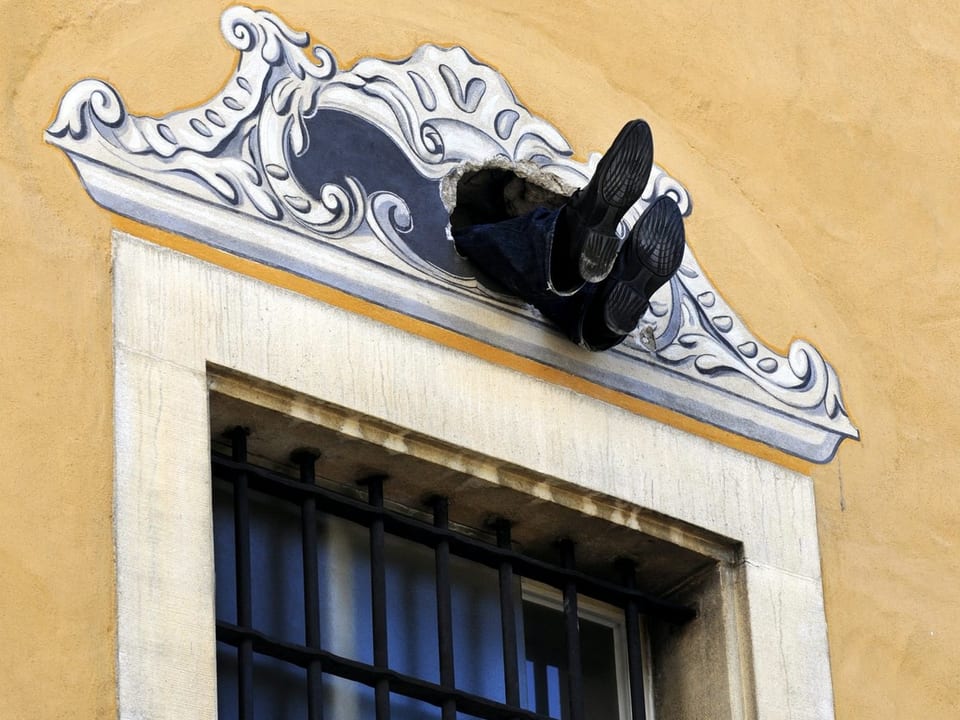 Zwei Füsse schauen aus einem Loch in der Fassade über einem Fenster heraus