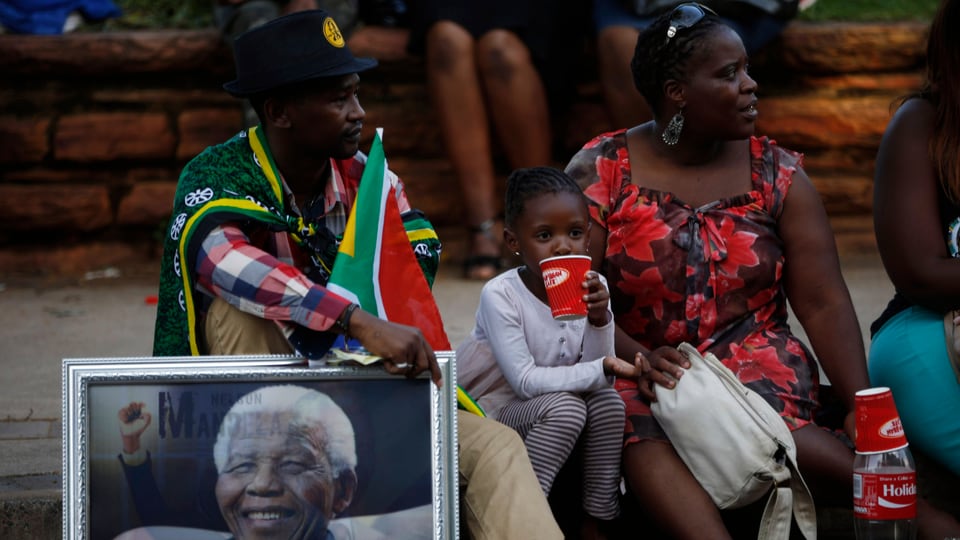 Eine Familie sitzt am Strassenrand, der Mann hält ein Bild Mandelas in der Hand.