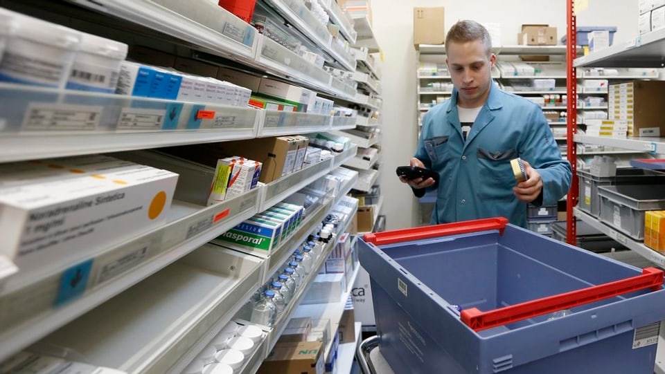 Ein Mann in blauem Kittel steht zwischen Regalen mit Medikamentenpackungen, ein blauer Plastikbehälter steht neben ihm auf einem Rollgestell.
