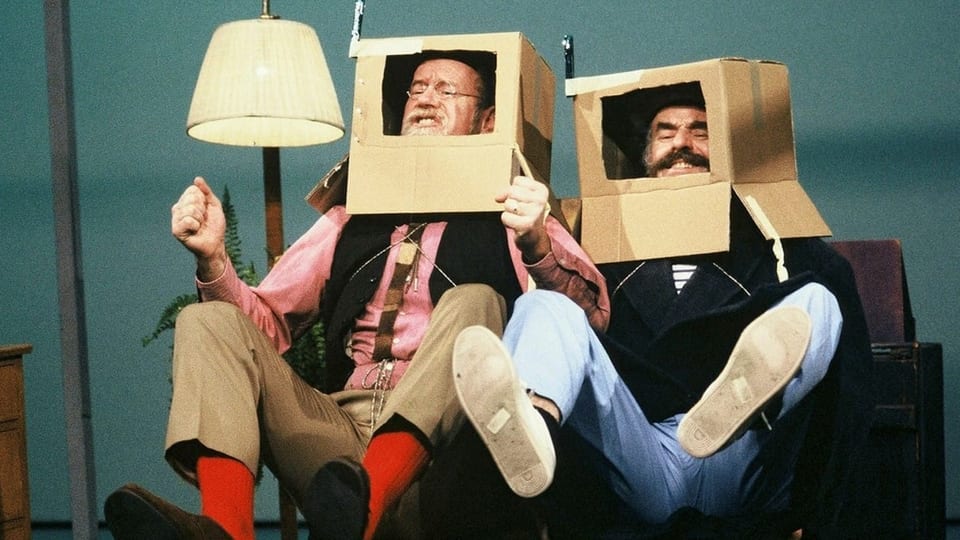 zwei Männer mit einem Karton über dem Kopf, beim Gesicht ein Quadrat ausgeschnitten, sitzen auf Sofa.