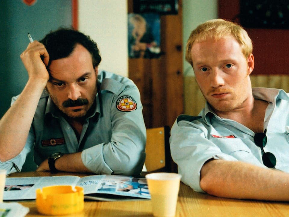 Filmszene: Brenner und Berti sitzen an einem Tisch, sie tragen die Uniformen von Krankenwagenfahrern.
