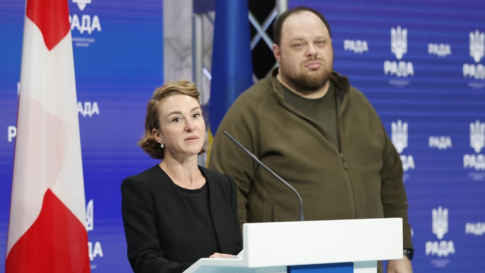 Frau an Rednerpult, daneben ein grosser Mann, Schweizer Fahne im Hintergrund