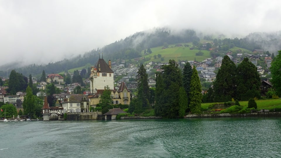 Schloss Oberhofen am grauen Thunersee, dahinter die Hügelzüge wolkenverhangen.